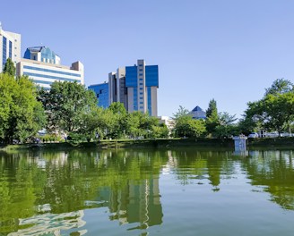 Modern buildings with blue-tinted glass windows rise behind a lush line of green trees. A tranquil pond in the foreground reflects the structures and vegetation, suggesting a harmony between urban development and nature.